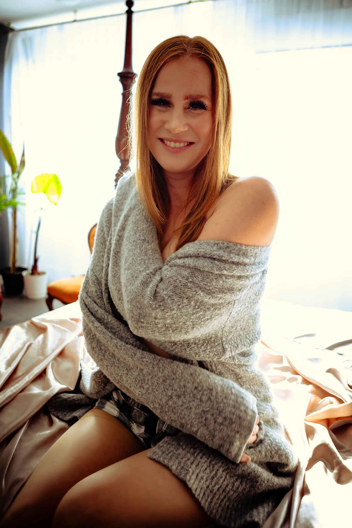 A woman seated by the studio window, smiling while wrapped in a soft grey blanket.